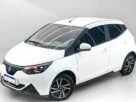 Visão em ângulo de três quartos da dianteira do JMEV Emova Easy, o carro elétrico mais barato do Brasil, um hatch compacto urbano com carroceria em dois tons (teto preto e pintura clara) posicionado em um estúdio fotográfico moderno.