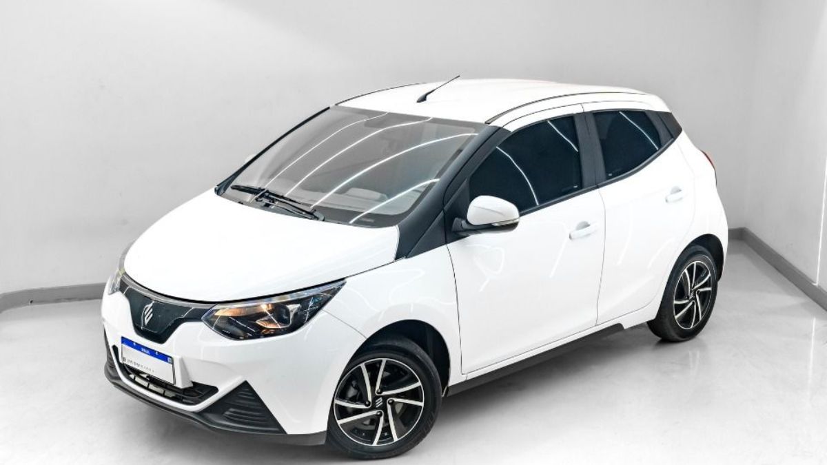 Visão em ângulo de três quartos da dianteira do JMEV Emova Easy, o carro elétrico mais barato do Brasil, um hatch compacto urbano com carroceria em dois tons (teto preto e pintura clara) posicionado em um estúdio fotográfico moderno.