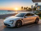 Fotografia de um carro esportivo elétrico Porsche Taycan branco estacionado e conectado a um carregador de parede (wallbox) em uma garagem litorânea. Ao fundo, destaca-se uma praia com coqueiros, um píer e o pôr do sol no horizonte, ilustrando o estilo de vida e a economia com o IPVA Carro Elétrico Ceará.
