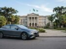 Fotografia de um sedã elétrico Mercedes-Benz cinza estacionado em uma via urbana no Paraná. Ao fundo, destaca-se um imponente palácio arquitetônico neoclássico cercado por árvores araucárias típicas da região, representando a transição para a mobilidade sustentável e as vantagens do IPVA Carro Elétrico Paraná.