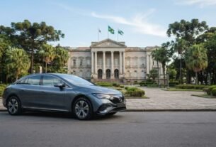 Fotografia de um sedã elétrico Mercedes-Benz cinza estacionado em uma via urbana no Paraná. Ao fundo, destaca-se um imponente palácio arquitetônico neoclássico cercado por árvores araucárias típicas da região, representando a transição para a mobilidade sustentável e as vantagens do IPVA Carro Elétrico Paraná.