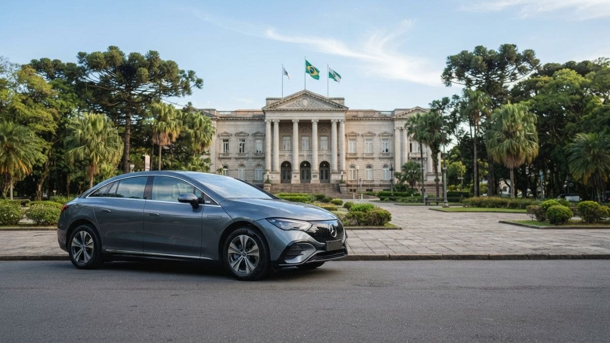 Fotografia de um sedã elétrico Mercedes-Benz cinza estacionado em uma via urbana no Paraná. Ao fundo, destaca-se um imponente palácio arquitetônico neoclássico cercado por árvores araucárias típicas da região, representando a transição para a mobilidade sustentável e as vantagens do IPVA Carro Elétrico Paraná.