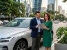 Fotografia de um homem de terno e uma mulher de vestido verde analisando informações em um tablet ao lado de um SUV híbrido prateado. Eles estão estacionados em uma avenida comercial moderna de São Paulo, ilustrando o planejamento do IPVA Carro Híbrido SP.