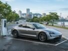 Fotografia de um Porsche Taycan cinza-prata estacionado em uma vaga de recarga na Orla do Guaíba, em Porto Alegre. O veículo está conectado a uma estação de carregamento rápida enquanto pedestres e ciclistas passeiam ao fundo, ilustrando o cenário da mobilidade e a dúvida se Carro Elétrico Paga IPVA no RS.