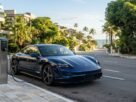Fotografia de um Porsche Taycan azul-marinho estacionado em uma rua de bairro nobre litorâneo, conectado a um totem de recarga na calçada. Ao fundo, uma ladeira arborizada leva em direção ao mar com coqueiros, ilustrando a mobilidade sustentável e os benefícios do IPVA Carro Elétrico Bahia.