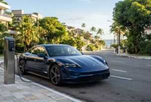 Fotografia de um Porsche Taycan azul-marinho estacionado em uma rua de bairro nobre litorâneo, conectado a um totem de recarga na calçada. Ao fundo, uma ladeira arborizada leva em direção ao mar com coqueiros, ilustrando a mobilidade sustentável e os benefícios do IPVA Carro Elétrico Bahia.