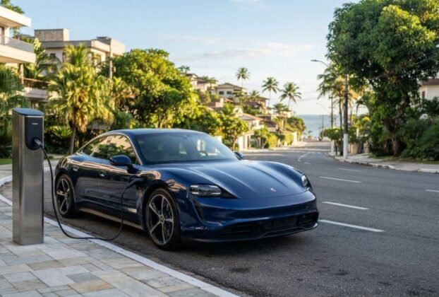 Fotografia de um Porsche Taycan azul-marinho estacionado em uma rua de bairro nobre litorâneo, conectado a um totem de recarga na calçada. Ao fundo, uma ladeira arborizada leva em direção ao mar com coqueiros, ilustrando a mobilidade sustentável e os benefícios do IPVA Carro Elétrico Bahia.
